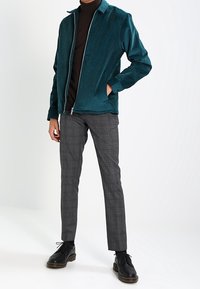 Turkusowa kurtka z korduroyu z zamkiem z przodu i spiczastym kołnierzem, zestawiona z szarymi spodniami w kratę i czarnymi butami na sznurówki.