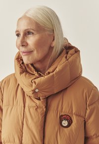 Siltā brūnā krāsā pūkains jaka ar lielu noliektu apkakli. Cīpsā ir izšūts lāča apdruka, un tai ir gluda, spīdīga tekstūra.