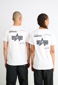 Valged puuvillased T-särgid, mille seljal on suur hall graafika, millel on tekst "ALPHA INDUSTRIES" ja kaks triibu mustrit. Mustad püksid on nähtavad all.
