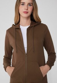 Sudadera con cremallera marrón de tela suave, con capucha ajustable, dos bolsillos frontales y puños ceñidos. Llevada sobre una camiseta blanca de cuello redondo.