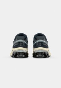 Baskets de sport en maille noire avec des accents beiges, présentant une semelle texturée avec un design segmenté et flexible, ainsi qu'un logo sur le côté.
