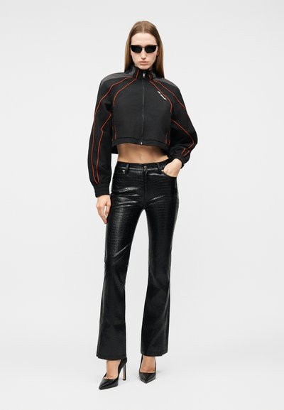 Γυναίκα που φοράει μαύρο κοντό μπουφάν με πορτοκαλί ρίγα, μαύρο δερμάτινο παντελόνι με υφή, μαύρες ψηλές γόβες και μαύρα γυαλιά ηλίου, στέκεται μπροστά σε λευκό φόντο.