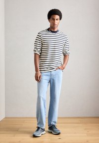 Homem com cabelo afro, vestindo uma camisa de manga longa listrada em preto e branco, jeans azul claro e tênis cinzas, em pé sobre um chão de madeira.