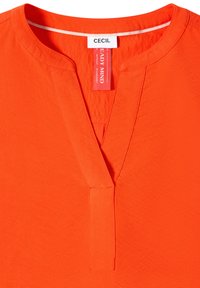 Haut orange vif à col en V, sans col, avec des étiquettes de marque visibles "CECIL" et "READY MIND" à l'intérieur de l'encolure.