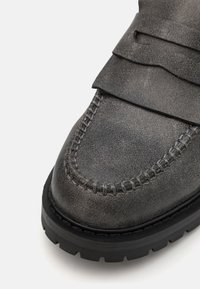 Šedá kožená bota s texturovaným povrchem, s prošívanou špičkou a silnou černou gumovou podrážkou pro lepší přilnavost. Mezi akcenty se nachází vrstvená krycí klapka.