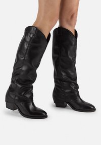Botas de cuero negro hasta la rodilla con diseño perforado, punta afilada y tacón bajo de bloque. Presentan un upper arrugado y detalles de costura.