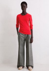 Czerwona bluzka z długim rękawem w połączeniu z szerokimi, szarymi spodniami o subtelnej fakturze. Modelka ma na nogach beżowe buty sportowe z czerwonymi akcentami.