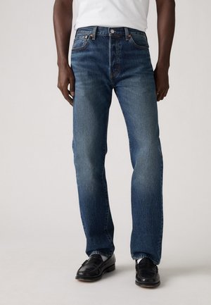 Dark blue denim jeans with a straight leg, featuring a traditional five-pocket design and brass hardware accents, worn with black shoes.