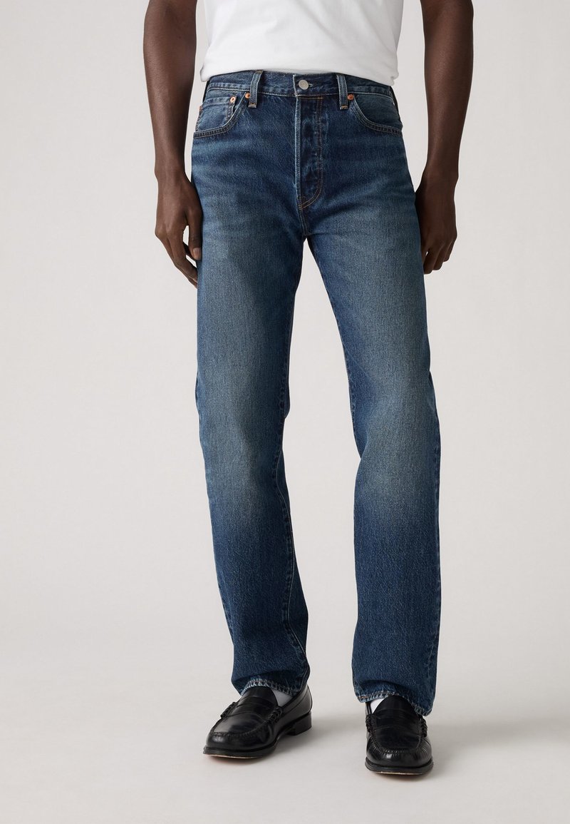 Jeans din denim albastru închis cu croială dreaptă, având un design tradițional cu cinci buzunare și accente din alamă, purtate cu pantofi negri.