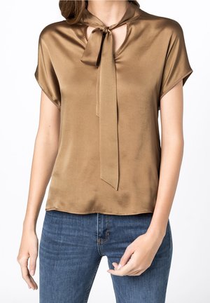 Mujer con una blusa de satén marrón de manga corta con un lazo atado en el cuello y jeans de mezclilla azul, con las manos relajadas a los costados.