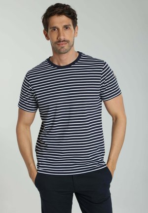 Homme portant une chemise à manches courtes rayée bleu marine et blanc et un pantalon sombre, debout les mains dans les poches devant un fond clair uni.