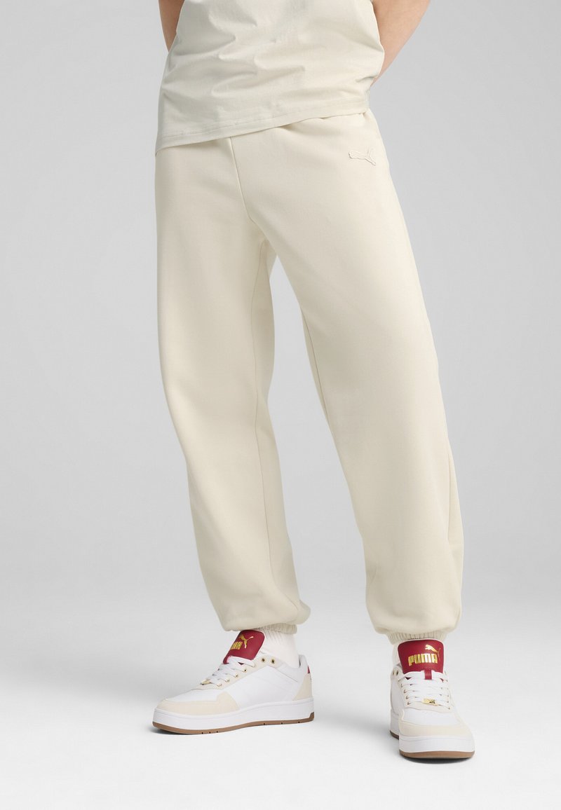 Cream-colored joggers with a smooth texture, featuring an embroidered logo on the front, paired with white sneakers and red accents.