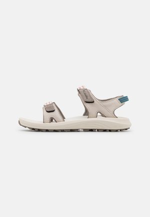 Sandalias de exterior beige con una suela blanca acolchada, correas ajustables y una parte superior texturizada. Presenta un acento azul vibrante en el talón.