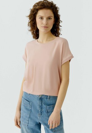 Femme aux cheveux bouclés châtain portant un haut rose pâle à manches courtes et un jean denim bleu avec de grandes poches avant, debout devant un fond uni.