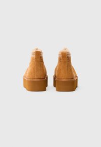 Un paio di stivali alla caviglia in suede beige con suole in gomma spessa e rivestimento morbido visibile, visti da dietro su uno sfondo chiaro e semplice.