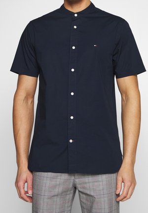 Mann, der ein marineblaues Kurzarmhemd mit Stehkragen und graue karierte Hose trägt, steht mit entspannten Armen.