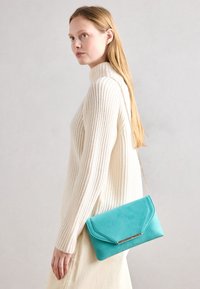 Türkise Clutch-Tasche mit umschlagbarem Design, goldener Kettenriemen und glatter Textur, getragen über einem cremefarbenen, gerippten Pullover.
