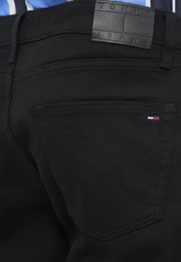 Mustmustest denim püksid nahkvarraste sildiga, millel on kiri "TOMMY JEANS." Sisaldab kahte tagumist taskut ja väikest triikolorist logodetaili.