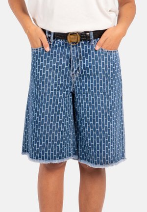 Person iført knelang blå denimshorts med et rektangulært mursteinsmønster, et svart belte med en rund messingspenne, hendene i lommene.