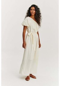 Krémové ľanové šaty uvoľneného strihu s naberanými rukávmi, opaskom na viazanie v páse a dlhou sukňou. Nosené s hnedými sandálmi.