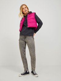 Gilet imbottito fucsia con bottoni a pressione, indossato sopra un hoodie nero. Abbinato a jeans skinny grigi e sneakers nere a basse. Sfondo semplice.