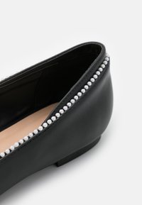 Chaussure plate en cuir noir avec un bout arrondi, présentant une rangée décorative de petites perles blanches le long du bord. Texture lisse, talon bas.