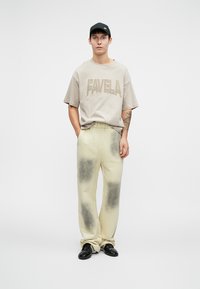 Béžové oversized tričko s nápisom "FAVELA", v kombinácii s off-white nohavicami s čiernymi tie-dye akcentmi. Čierna čiapka a lesklé čierne topánky.