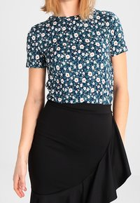 Květinový top s krátkým rukávem na námořnickém pozadí, zdobený bílými sedmikráskami a zelenými listy, doplněný černou zavinovací sukní s texturovaným povrchem.