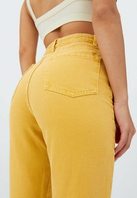 Kvinna i senapsgula högmidjade jeans och en beige croppad ribbad topp, sedd bakifrån från midjan och ner.