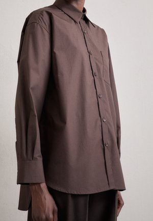 Mann trägt ein locker sitzendes, langärmeliges, braunes Hemd mit Knöpfen und einer Brusttasche vor einem schlichten hellen Hintergrund.