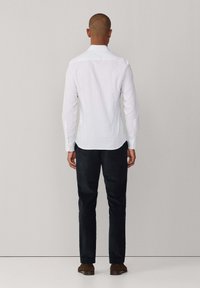 Witte langemouwen overhemd met een subtiele textuur en een traditionele kraag, gecombineerd met donkere broek en bruine schoenen, gezien van achteren.