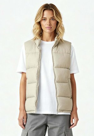 Blonde Frau trägt eine beige gesteppte Weste über einem weißen T-Shirt und grauen Cargo-Hosen, steht vor einem schlichten hellen Hintergrund.