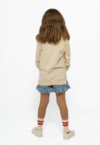 Beige långärmad topp, blå rutig volangkjol, vita skor och randiga strumpor. Håret är brunt och vågit. Bakifrån.