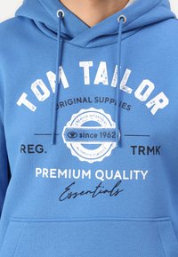 Blauer Hoodie aus Baumwolle mit einer Kordelzugkapuze. Er hat weißen und schwarzen Text, darunter "Tom Tailor" sowie dekorative Logodetails.