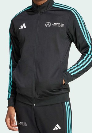 Osoba ubrana w czarny dres AMG Petronas z turkusowymi paskami i białymi logo, ręce w kieszeniach, stojąca na jednolitym tle.