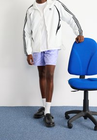 Vit jacka med dragkedja och svarta ränder, lila mönstrade shorts, svarta skor med dekorativa detaljer och vita strumpor. Blå kontorsstol i närheten.