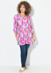 Tunika dai colori vivaci con modelli astratti in rosa, blu e oro, con scollatura a barca e maniche a tre quarti. Abbinata a jeans scuri.