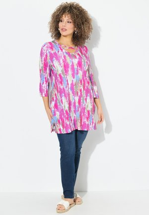 Helle, farbenfrohe Tunika mit abstrakten Mustern in Pink, Blau und Gold, einem Rundhalsausschnitt und dreiviertellangen Ärmeln. Kombiniert mit dunklen Jeans.