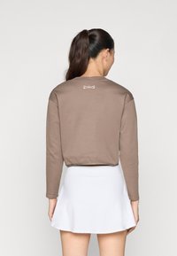 Καφέ μπλούζα με μακρύ μανίκι και στρογγυλή λαιμόκοψη, με λευκή λεπτομέρεια λογότυπου στο πίσω μέρος, σε συνδυασμό με λευκή φούστα σε αέρινη γραμμή. Λείος υφασμάτινος τύπος.