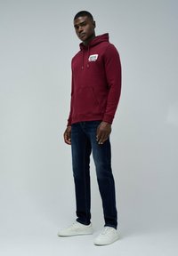 Młody mężczyzna ubrany w bordowy hoodie z logo "Salsa Jeans", ciemnoniebieskie dżinsy i białe trampki, stojący na tle jasnego, jednolitego tła.