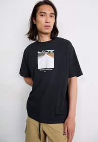 Svart kortermet t-skjorte med grafisk trykk av en vei og spiraldesign, med tekst "Det finnes ingen mållinje" og Nike-logo.