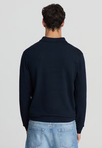 Polo shirt navy a maniche lunghe con colletto e polsini a costine, caratterizzato da una texture morbida e una vestibilità rilassata, indossato sopra jeans azzurri chiari.