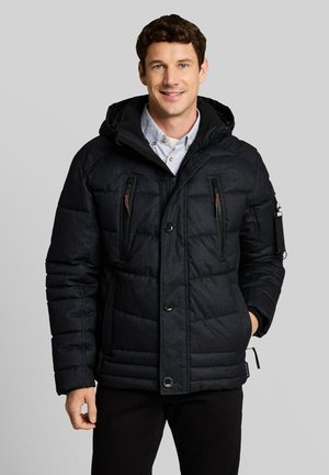 Uomo che indossa una giacca invernale imbottita nera con cappuccio sopra una camicia azzurra con bottoni, in piedi davanti a uno sfondo grigio chiaro.