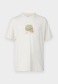 Camiseta de algodón color crema con un corte relajado; presenta un gráfico de una calavera y un diseño de rejilla en verde y rojo. Mangas cortas y cuello redondo.