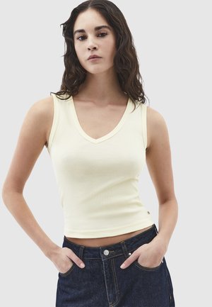 Jonge vrouw met lang donker golvend haar, gekleed in een mouwloos lichtgeel V-hals topje en donkerblauwe spijkerbroek, met handen in de zakken.