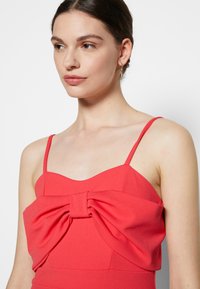 Vestido coral com alças finas, apresentando um grande laço na parte da frente. A textura suave e o design estruturado destacam a silhueta.