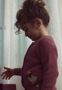 Pleteni roza cardigan z teksturno vzorcem, ki ima vezenino race z mašno na levi strani. Model je otrok s kodrastimi lasmi.