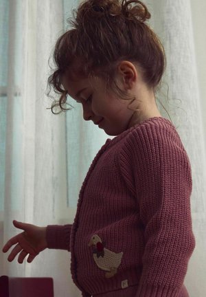 Gebreide roze cardigan met een gestructureerd patroon, voorzien van een eendenborduring met een strik aan de linkerkant. Het model is een kind met krullend haar.
