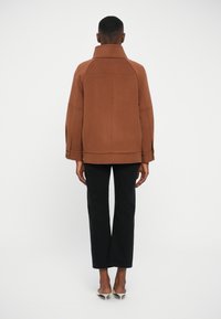 Giacca in tessuto marrone con colletto alto e maniche larghe, caratterizzata da cuciture distintive e dettagli con bottoni, abbinata a pantaloni neri.