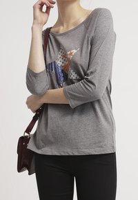 Grauer Langarmoberteil mit lockerer Passform, mit einem Sternendesign, das mit blauen, orangefarbenen und weißen Pailletten verziert ist. Schwarze Hosen und eine burgunderfarbene Tasche.